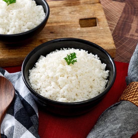 A bowl of cooked white rice with parsley sits on a wooden table with a checkered cloth and wooden spoon nearby. In the background, theres another bowl of rice next to an American Reserves Protein Bucket, known for its nutritious freeze-dried proteins and an impressive 25-year shelf life.