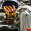 A pan with sausages sizzles on a portable camping stove, while the Kelly Kettle® Ultimate Scout Kit by American Reserves sits nearby, outdoors by a stream.