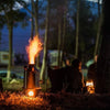 At dusk in a wooded area, flames flicker from a metal camping stove, casting silhouettes of figures and lanterns. Nearby, the Kelly Kettle® Ultimate Scout Kit by American Reserves is ready to brew warmth as night envelops the campsite.