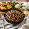 A black bowl containing dry dog food pellets sits on a patterned cloth. Behind it is a bowl of American Reserves Protein Bucket, featuring cooked vegetables, meat, and freeze-dried proteins. Potatoes and rosemary sprigs are nearby, with a fork resting on the cloth.