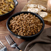 A black bowl of American Reserves Protein Bucket freeze-dried ground beef sits on a wooden surface. Sliced mushrooms lie on a cutting board in the background, flanked by two forks resting on a brown cloth. A bowl with pasta and mushrooms peeks into view.