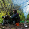 A person sits on a green chair in the woods, dressed in outdoor clothing and boots. Nearby, an American Reserves Kelly Kettle® Ultimate Base Camp Kit emits steam over a small campfire, illustrating essential base camp gear.