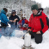 People in winter clothing gather around a campfire in a snowy forest, as one person tends to an American Reserves Kelly Kettle® Ultimate Base Camp Kit.